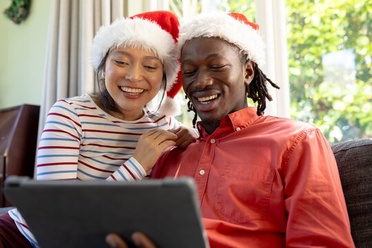 Happy diverse couple in christmas hats having tablet video call smiling in sunny living room