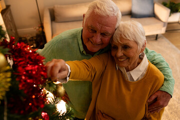 Happy caucasian senior couple embracing and decorating christmas tree in living room at home