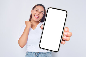 Fototapeta na wymiar Beautiful excited Asian girl wearing casual white t-shirt showing mobile phone with blank white screen, raising fist, celebrating good luck isolated on white background. People lifestyle concept