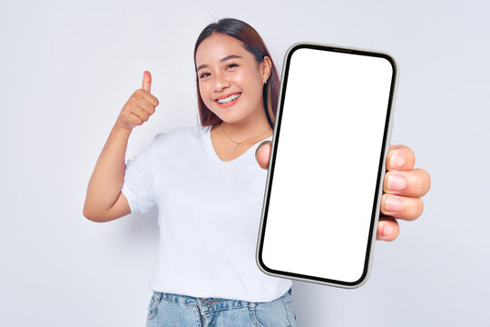Beautiful cheerful Asian girl wearing casual white t-shirt showing mobile phone with blank black screen and raising thumb up isolated on white background. People lifestyle concept