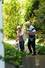 Fototapeta premium Happy caucasian senior couple walking with flowers and watering can in sunny garden