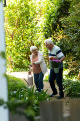 Happy caucasian senior couple walking with flowers and watering can in sunny garden