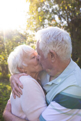Happy caucasian senior couple embracing and kissing in sunny garden, copy space