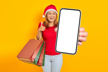 Smiling young Asian woman in Santa Claus hat holding blank screen phone and shopping bag while...