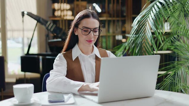 Focused Business Woman In Eyeglasses Typing At Modern Laptop In Cafe Interior Attractive Young Girl Manager Looking Computer Screen Sitting In Restaurant Serious Woman Company Worker Surfing Internet.