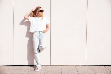 Portrait of beautiful smiling model in sunglasses. Female dressed in summer hipster white T-shirt and jeans. Posing near wall in the street. Funny and positive woman having fun outdoors