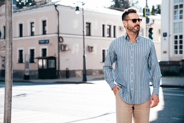 Fototapeta premium Portrait of handsome confident stylish hipster lambersexual model. Sexy modern man dressed in blue shirt and trousers. Fashion male posing on street background in Europe city at sunset. In sunglasses