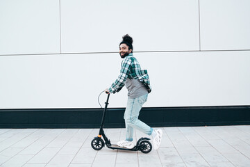 Trendy smiling bearded man in casual clothes riding electric scooter in urban background. Handsome model posing in the street near wall. Hipster guy with curly hairstyle