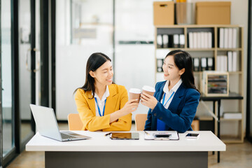 Happy two Asian business woman holding coffee cup in coworking office in morning light.