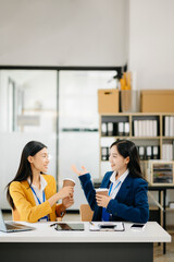Happy two Asian business woman holding coffee cup in coworking office in morning light.