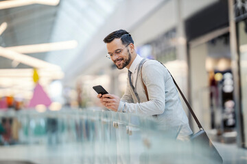 A happy young man is standing at the shopping mall and checking on discounts on the phone.