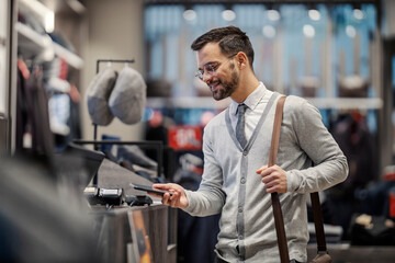 A happy young elegant man is using phone app in a boutique for paying bill.