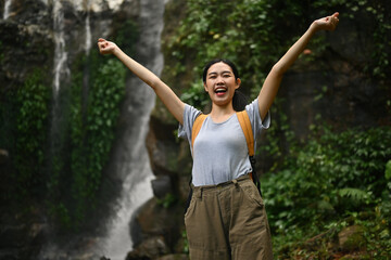 Cheerful female traveler with backpack raising arms enjoying the view of beautiful tropical waterfall