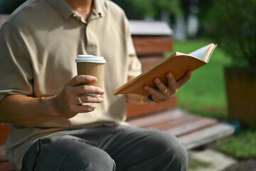 Middle aged and drinking coffee and reading book while sitting on bench at summer park