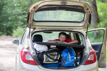 A child on a journey. A boy looks out of the trunk of a car filled with huge bags.