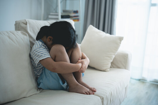 Asian kid girl upset lonely bullied little Sitting hugging your knees on the sofa couch in living room at home feels abandoned abused, sad alone, charity adoption concept.