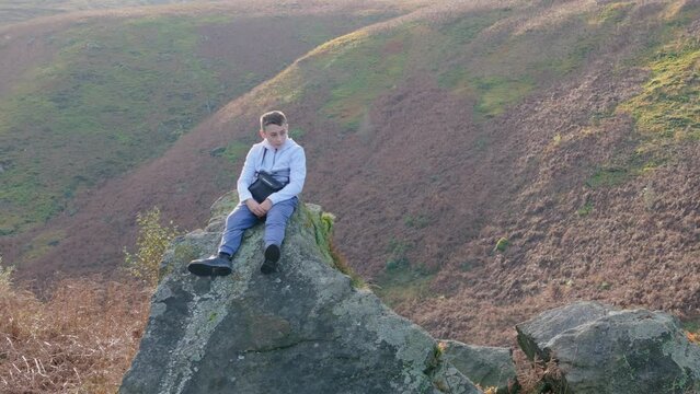 Aerial footage circles a pensive young boy atop a substantial rock outcrop in the moorland, lost in thought.