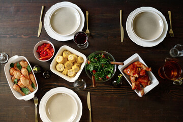 Top view of wooden table with homemade roasted and baked poultry, vegetable salad, corn and fresh red pepper, plates and silverware