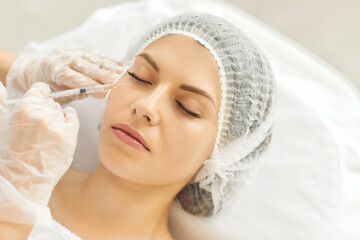 Young lady receives filler injection in lower eye lid area done by specialist at beauty clinic. Closeup woman's head in medical cap, hands in gloves holding syringe. Aesthetic facial treatment concept
