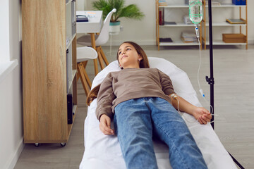 Child getting intravenous vitamin or medication therapy at clinic or hospital. Calm little girl lying on bed and receiving medical infusion through IV line. Medicine, health care, treatment concept - obrazy, fototapety, plakaty