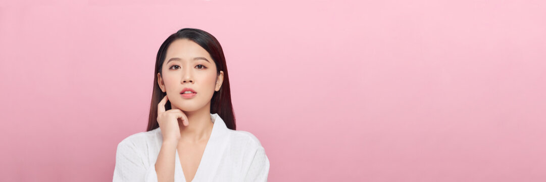 Beautiful asian girl model with natural makeup touching glowing hydrated skin on pink background, panorama