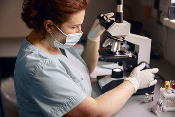 Middle aged lab technician adjusts microscope to research material sample at workplace