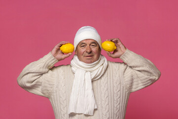 A man in a white sweater and hat on a pink background holds two lemons.