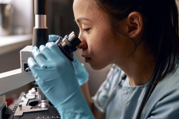Asian woman in blue gloves looks into microscope lens sitting at workplace in laboratory
