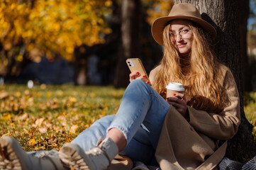 Beautiful blonde woman in a coat and hat sits on a plaid under a tree in an autumn park on a sunny...