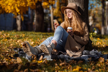 Beautiful blonde woman in a coat and hat sits on a plaid under a tree in an autumn park on a sunny day