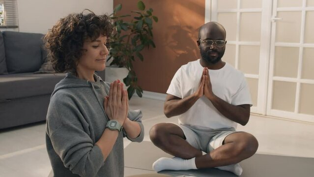 Tilt up shot of multi-ethnic couple sitting on floor with mats and different sport equipment watching yoga tutorial on black laptop