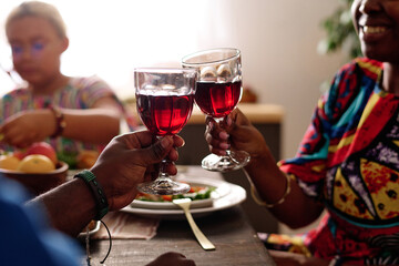 Hands of young African American couple with red homemade wine clinking with wineglasses while celebrating kwanzaa by served table
