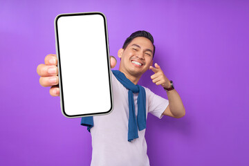 Cheerful young Asian man pointing index finger at empty smartphone screen isolated on purple background. Advertising new mobile app, mockup concept - obrazy, fototapety, plakaty