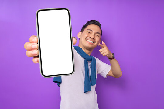 Cheerful young Asian man pointing index finger at empty smartphone screen isolated on purple background. Advertising new mobile app, mockup concept