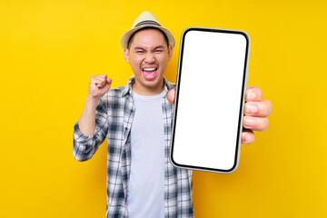 excited handsome Asian young man 20s wearing casual clothes hat Showing mobile cell phone with blank screen and making winner gesture isolated on yellow background. People lifestyle concept - obrazy, fototapety, plakaty