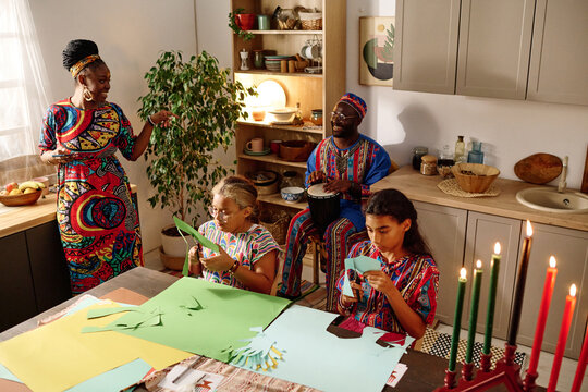 Two creative African American girls in national attire cutting paper while preparing greeting cards for kwanzaa holiday against their parents