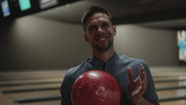 Portrait of caucasian middle aged man holding pearl bowling ball and smiling. Player in dark bowling club with lanes at back rejoices before throwing heavy ball.