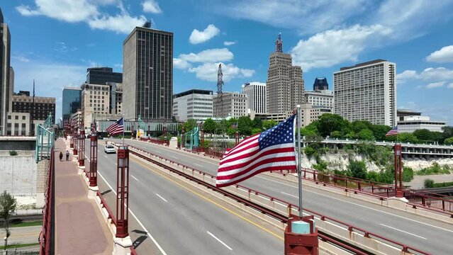Saint Paul, Minnesota skyline on a sunny day, showcasing tall buildings, a bridge with American flags, and a riverfront beneath a clear blue sky.