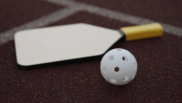 White plastic pickleball ball with holes lies near racket closeup on sports field. Shot of sports equipment on street outdoors after match on rubber surface with markings.