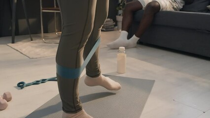 Close-up shot of unrecognizable woman standing on mat in sport clothes using mini band and African American man sitting on couch in background