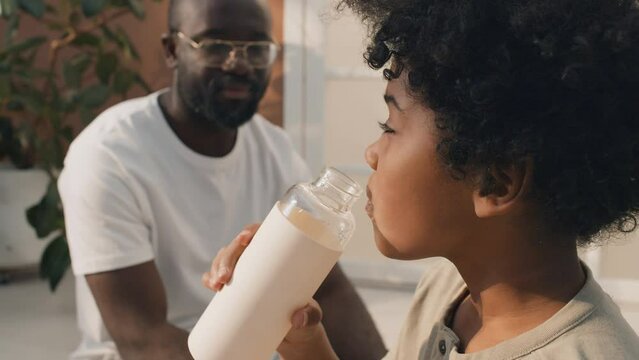 Medium close-up shot of young boy with curly hair drinking water while his father on background explaining importance of staying hydrated