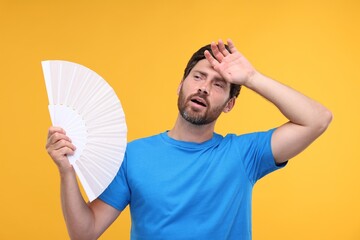 Unhappy man with hand fan suffering from heat on orange background - obrazy, fototapety, plakaty