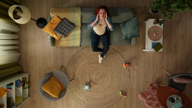 In the shot from above, a pregnant woman sits on a sofa in a room and holds her head. She has a headache, she is irritated, sad. Childrens toys lie on the floor nearby.