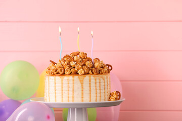Caramel drip cake decorated with popcorn and pretzels near balloons against pink wooden background,...