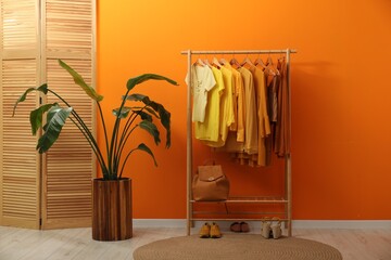 Rack with different stylish women`s clothes, shoes, backpack and green houseplant near orange wall indoors