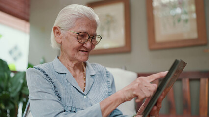 Happy Asian senior woman looking through a family photo album while leisure time at home. Elderly older grandmother looking at family photographs. Important moments and memory of life - obrazy, fototapety, plakaty