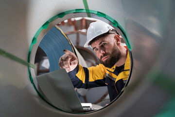 Male factory worker using laptop computer inspecting quality rolls of galvanized or metal sheet in...