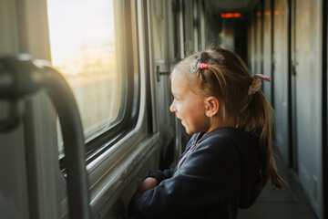 Toddler child looking through train window on sunset, bright sunlight, atmospheric travel by railway with kids. Girl happy exploring the way in evening. Exciting family trip - obrazy, fototapety, plakaty
