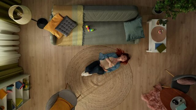 In the shot above, a pregnant woman in the room is lying on the floor. She is holding her stomach, she is in a lot of pain, squirming in pain. A couch with childrens toys stands nearby.