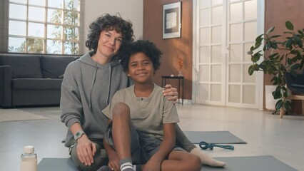 Portrait of mother and her adopted son sitting in their living room on mats with sport equipment looking at camera and smiling